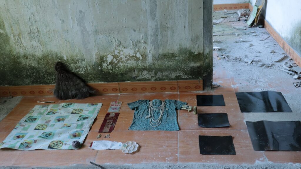 Action-installation éphémère de Sarah Venturi, extrait de la série Il était une fois (mine de Wumushung, 2024). Objets trouvés (balai traditionnel, morceau de toile cirée, emballages, calligrammes en plastique doré, fragment de calendrier, palette, billes, vêtement bleu imprimé prolongé par un fragment d'avion jouet imitant une prothèse de bras, lien de store simulant un collier de perles, radiographies médicales) alignés au sol (Meitanba, Chine).